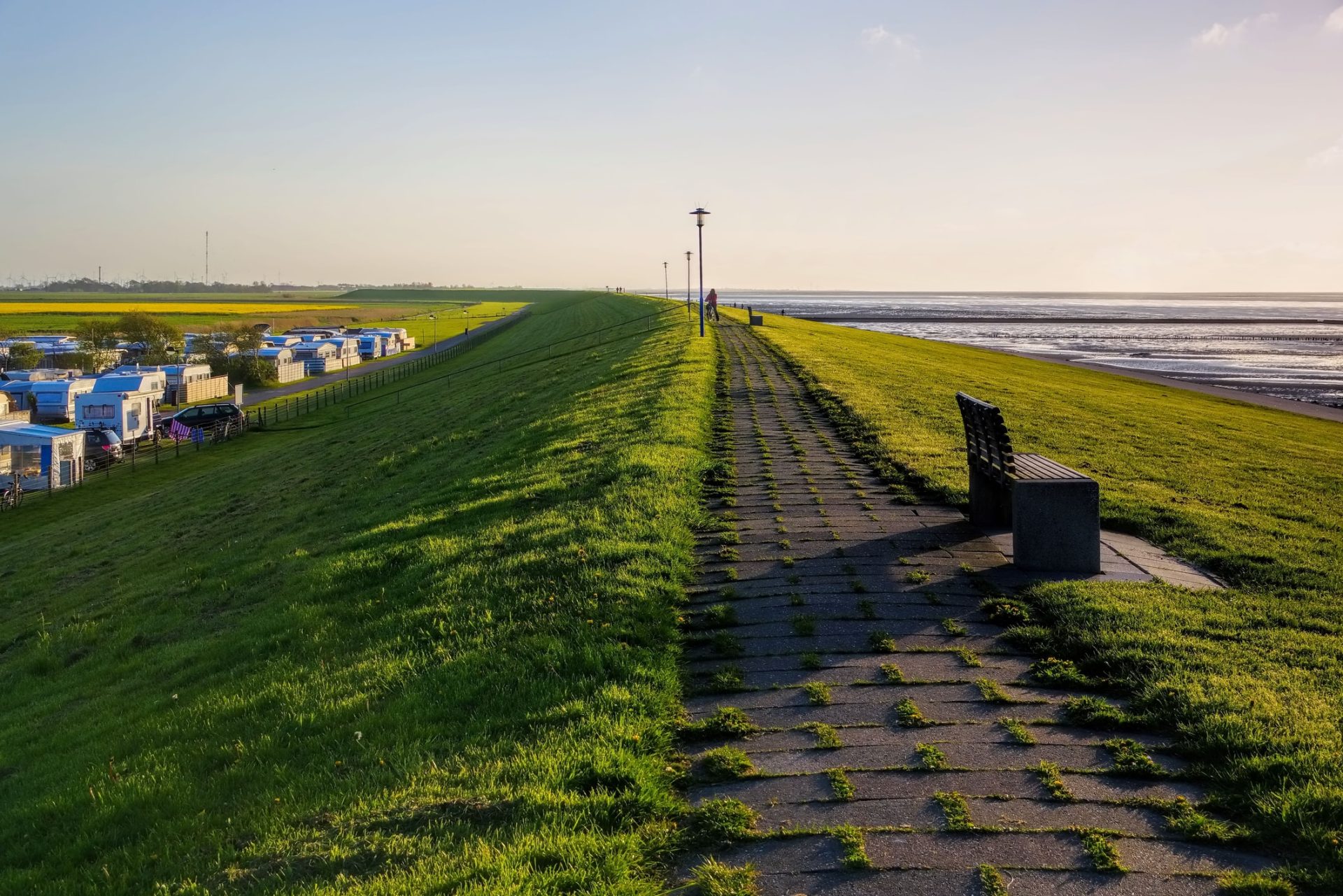 Gepflasterter Weg auf einem grasbewachsenen Deich entlang der Küste bei Sonnenuntergang. Rechts ist die Nordsee bei Ebbe zu sehen, links unten stehen zahlreiche Wohnwagen und Camper auf einem Campingplatz. Ein einzelner Holzbänke steht am Weg mit Blick auf das Meer. Die Szene wirkt ruhig und idyllisch.