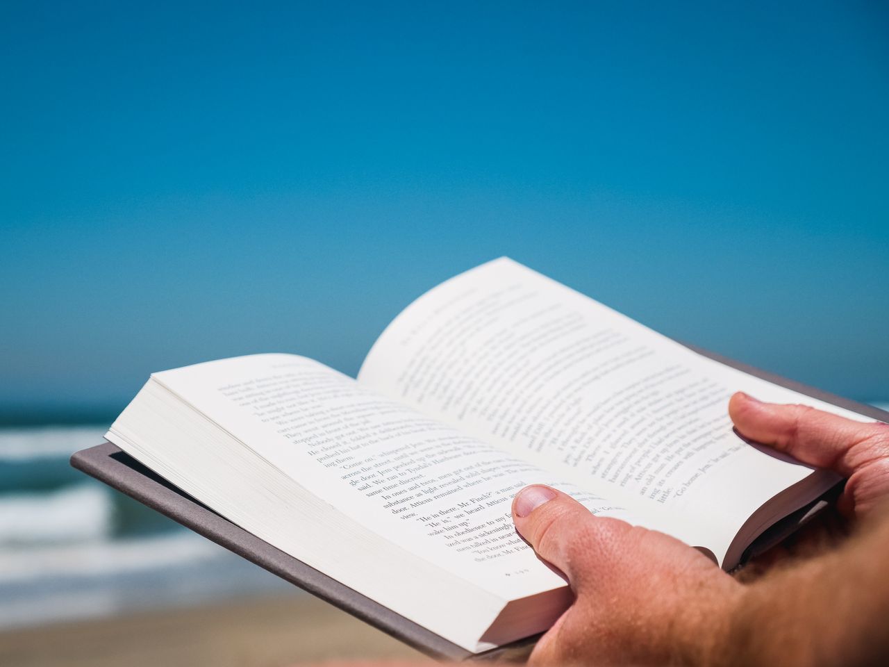 Nahaufnahme von Händen, die ein Buch am Strand mit Ozean und klarem Himmel halten.